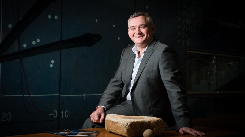 Greg Pratt seated on a desk and dressed in a suit.