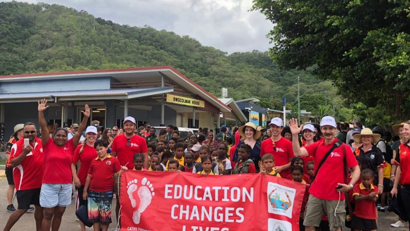 A group of men, women and children in red school polo shirt with a crowd of Indigenous children behind red banner with white writing on banner ‘Education Changes Lives.’