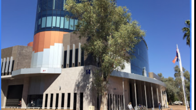 Blue, orange, white and beige building with tree in front and footpath with pavers in front of that. Aboriginal flag at right.