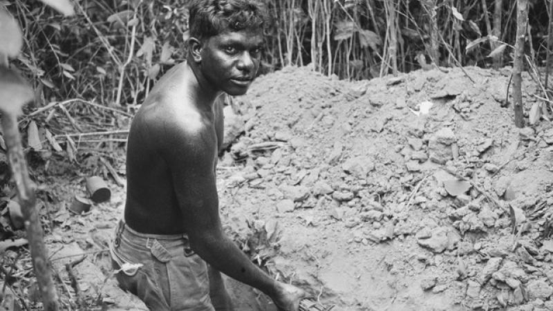 A young man wearing shorts but bare chested holds a shovel in his hand while he stands in a trench. In front of him is a pile of soil and beyond that is thick foliage.
