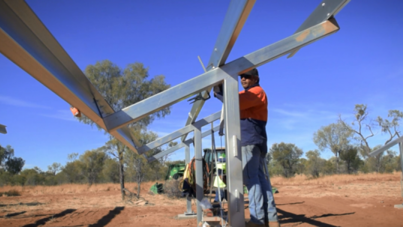 Aboriginal man dressed in workwear, hat and sunglasses works on a steel structure set on red earth with grass and trees in the background and a bright blue sky overhead.