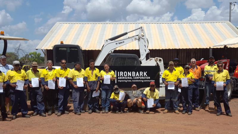 A group of men and women holding certificates while standing in front of heavy machinery and a shed.