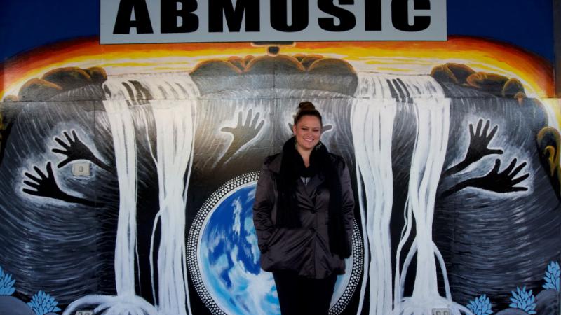 Indigenous woman standing in front of a large grey and white and blue painting of black hands and white waterfalls and the word ABMUSIC at the top.