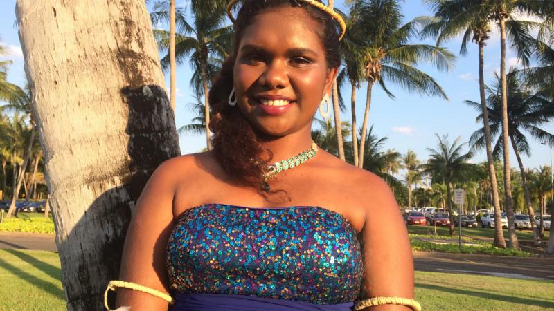 Young Tiwi Island woman in purple dress and headdress standing in front of a palm tree.