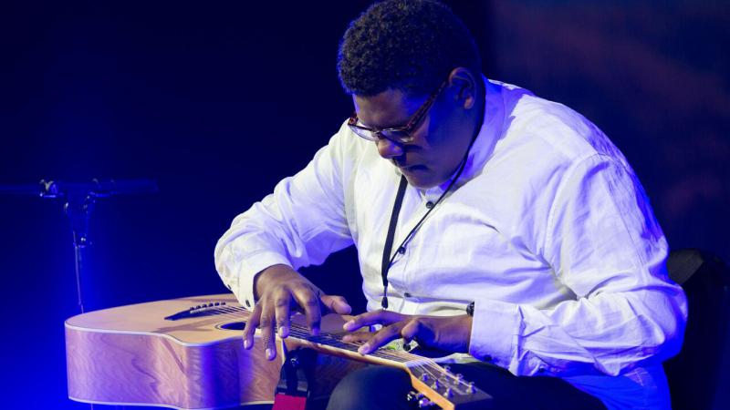 Indigenous man playing a guitar and wearing pale shirt and dark trousers.