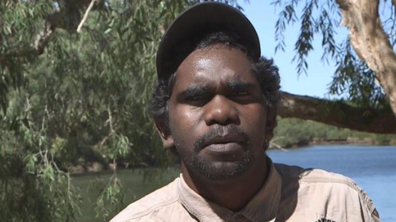 Aboriginal young man dressed in ranger uniform in foreground with river and trees in the background.