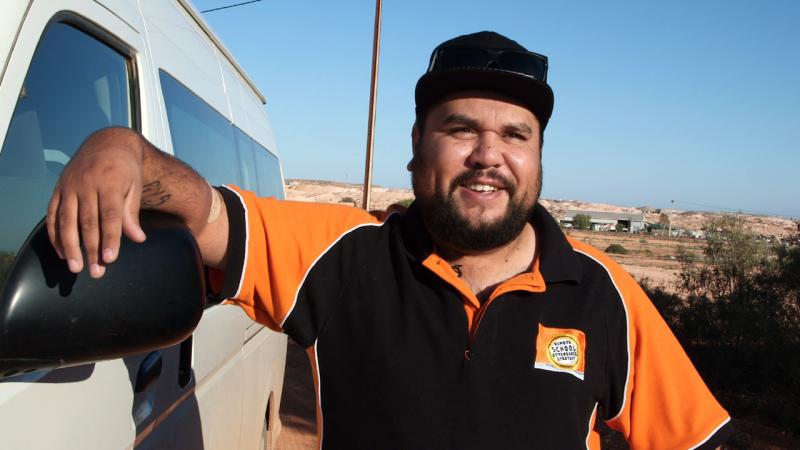 Indigenous man leaning up against a white van with buildings in the background.