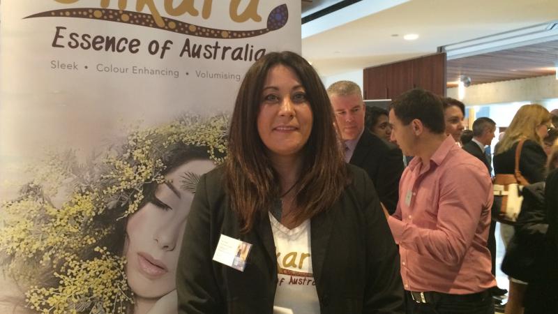 A woman, Julie Okley, stands in front of a poster displaying the words Dilkara, Essence of Australia. There are other people in the background.