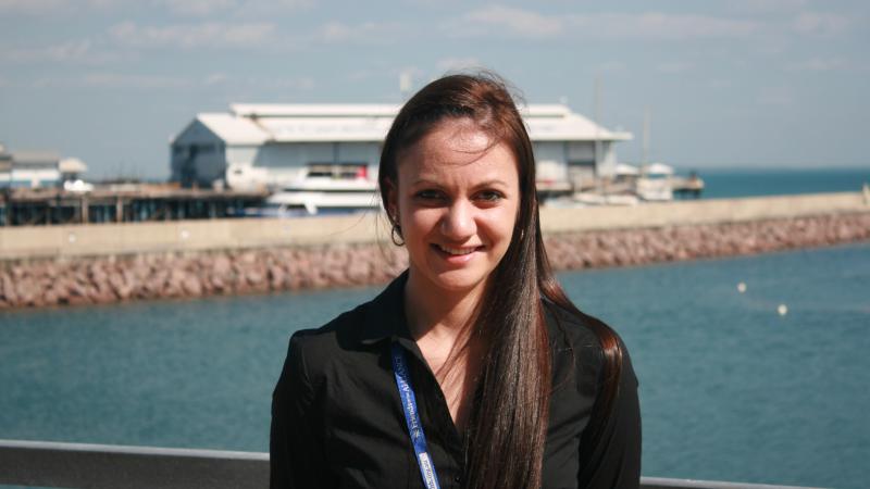 Woman dressed in black shirt stands in foreground with blue water and buildings in the background.