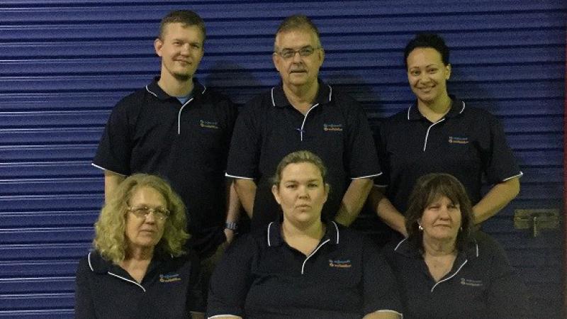 Two rows of people dressed in blue polo neck shirts in front of a blue roller door; three standing in the back row and three are sitting in the front.