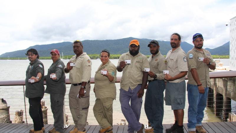 Line of Indigenous men and women dressed in ranger uniforms display their individual ranger accreditation. In the background is a wharf, a bay and a hill.