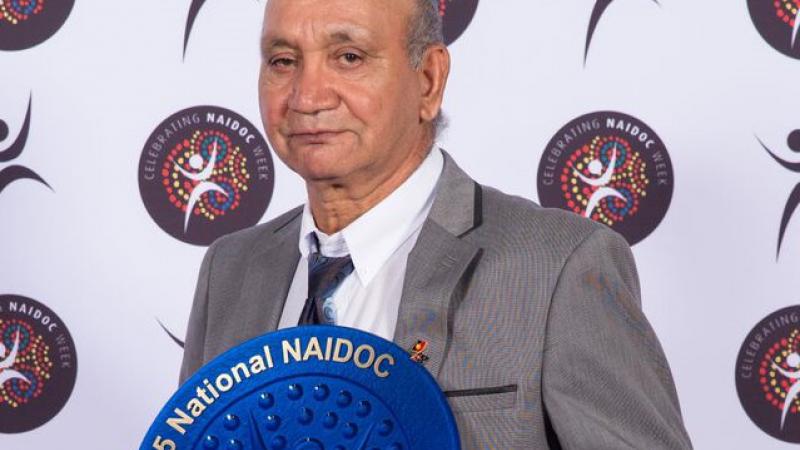 Indigenous man dressed in grey suit holding blue plate displaying the text 2015 National NAIDOC