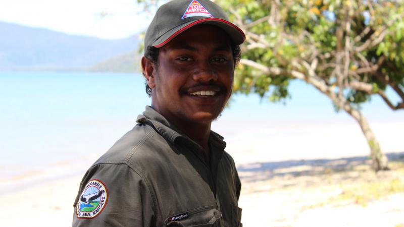 Indigenous young man dressed in ranger clothing standing near tree with body of water and hills in the background.