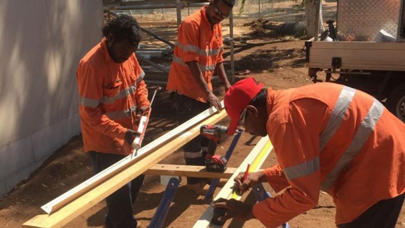 Three Aboriginal men dressed in orange work clothing and standing on red dusty soil next to a building prepare timber for use in construction.