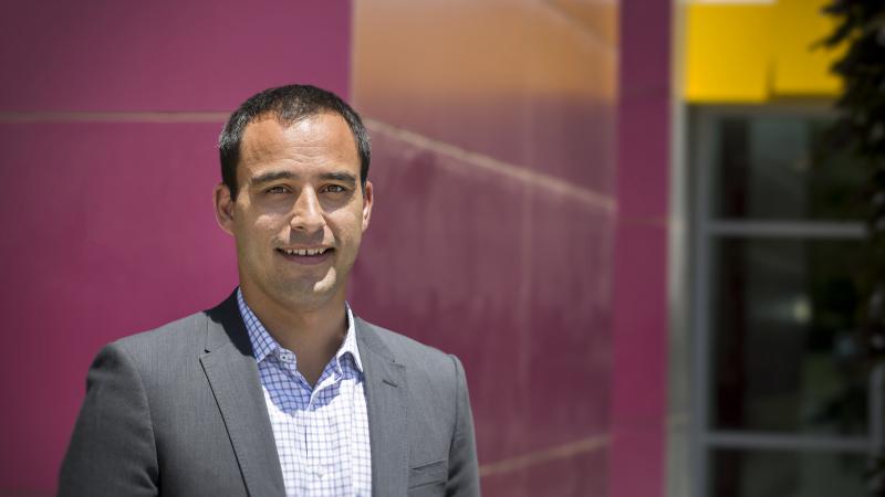 Man in grey suit standing in front of a maroon wall.