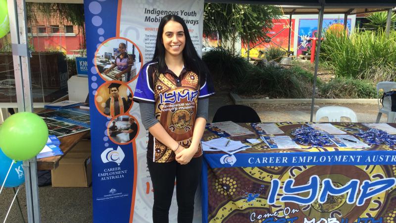 Young Indigenous woman standing in front of sign and to the side of a display table for Indigenous Youth Mobility Pathways. She is dressed in black pants and a colourful shirt showing Indigenous designs.