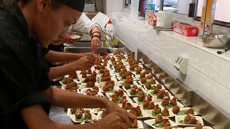 Indigenous woman dressed in black prepares multiple food dishes on stainless steel bench with fellow chefs in the background.