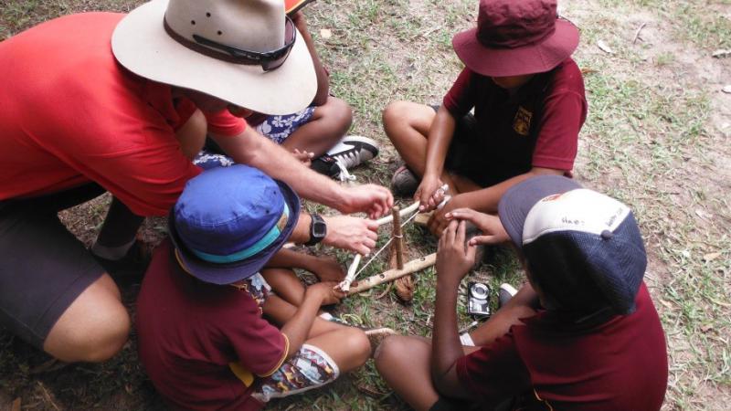 Man in hat teaches four Indigenous students in hats and maroon shirts how to start a fire