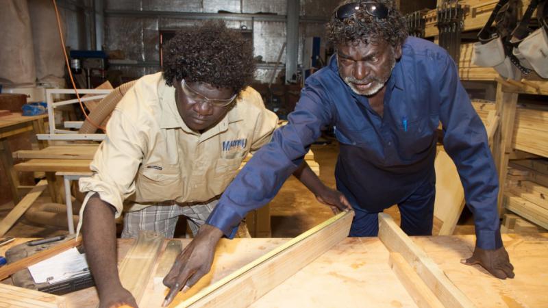 Two Indigenous men working at a wood working bench with the elder man directing the younger