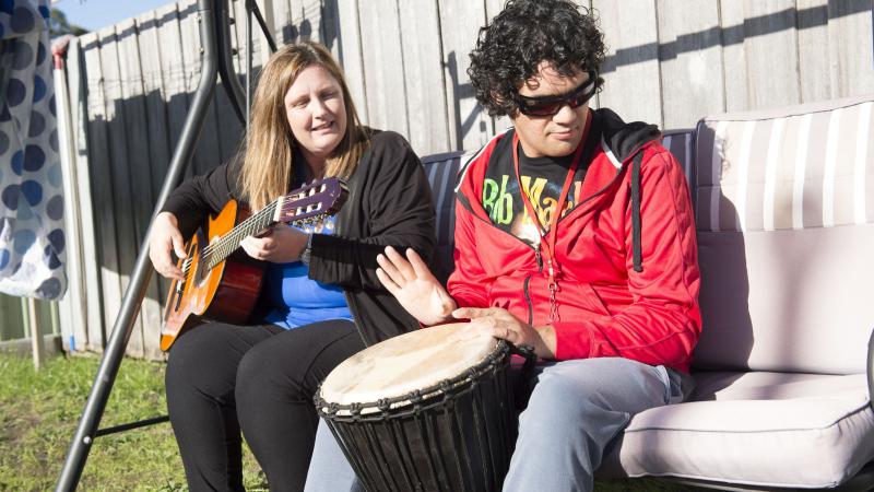 Seated on a large swing chair, an Indigenous woman dressed in black plays a guitar while an Indigenous man in red top and grey pants plays a large bongo drum.