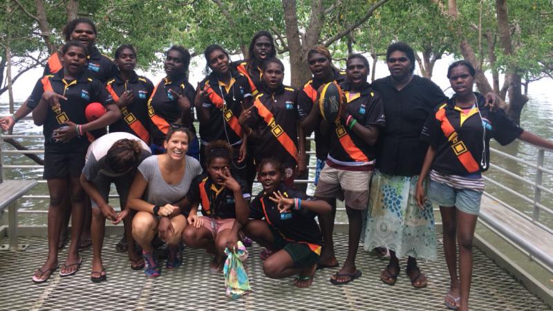 A group of young Indigenous women and their female supervisor standing near a body of water and trees.