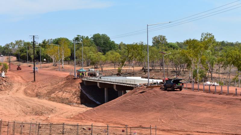 In the foreground is ochre coloured soil behind which is a concrete bridge with vehicles nearby and in the background are trees and blue sky.