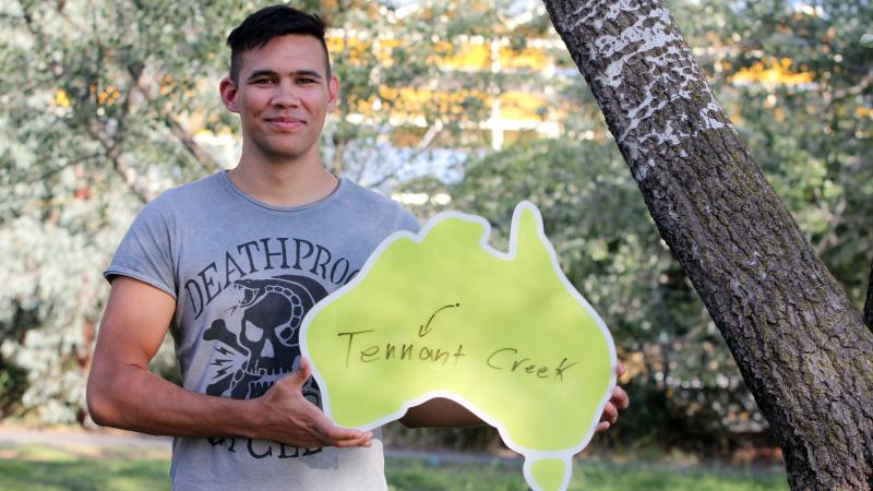 Young Indigenous man wearing grey t-shirt standing beneath a tree and holding a cardboard cut-out of Australia featuring a dot with an arrow to the words Tennant Creek.