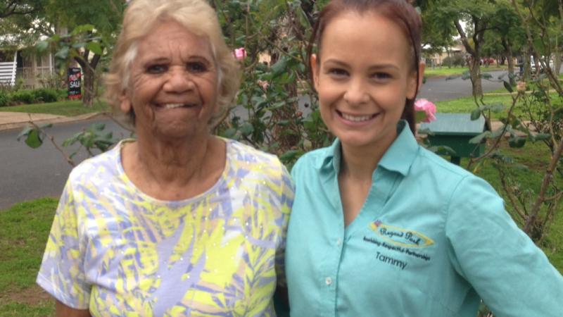 Elderly Indigenous woman in yellow and grey dress stands with arm around young Indigenous woman dressed in aqua coloured shirt in front of a flowering bush in a suburban street.