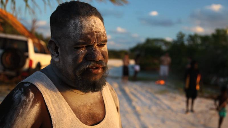 Aboriginal man Timmy Burarrwanga in foreground with others in background outdoors, standing on sandy soil.