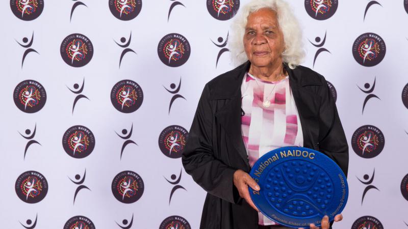 Elderly Indigenous woman, dressed in dark brown over coat and pink and white blouse, holding a blue award in the shape of a plate.