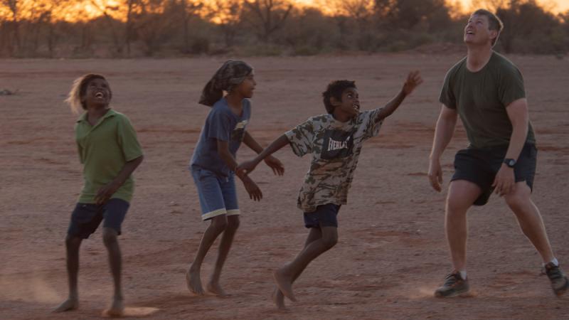 Three Aboriginal children and a non-Aboriginal man look upward on a dusty field with trees in the background.