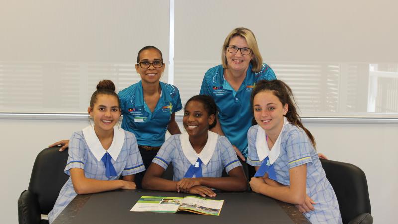 Two women in aqua coloured shirts stand behind three young women in pale blue school uniforms seated at a table.