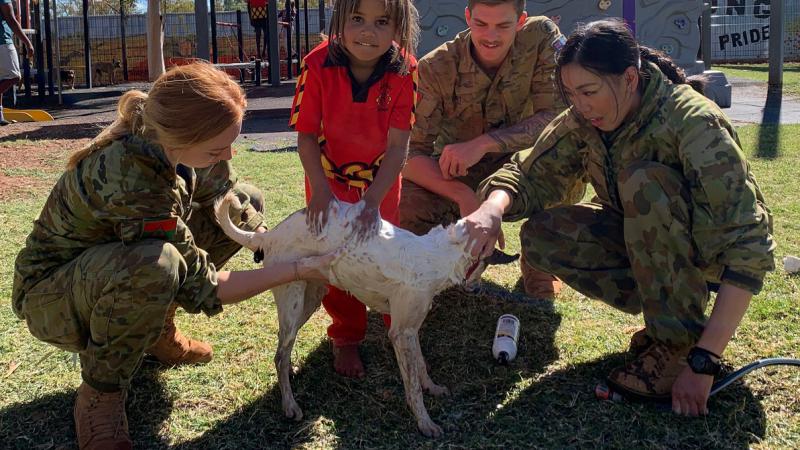 Young Aboriginal girl in red school uniform and three soldiers (2 women and 1 man) surround a white dog on a grassed area. One soldier holds a hose. In the background is a playground.