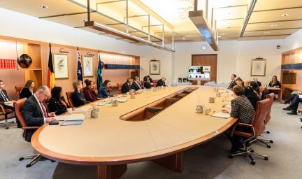 Group of people formally dressed sit around a board table in a large room. In the background is wood pannelling, more people, a door, flags and other office content.