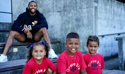 A man in black top and shorts sits on a step in an outdoor area. Three children in red tops sit below him.