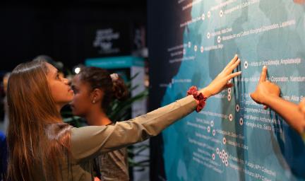 Young woman with long brown hair touches a map of Australia featuring place markers and names. In the background is another young woman with her hair in a bun. Beyond her is a display. In the foreground is an arm touching the map.