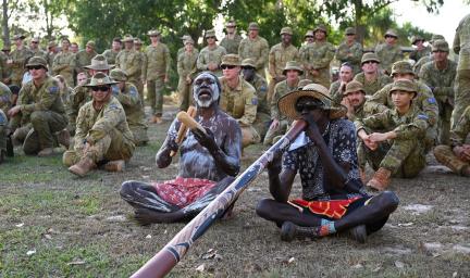 Two men in traditional paint and wear sit on the ground. One blows into a didgeridoo while the other bangs sticks together. Behind them are men and women dressed in army wear. Behind them are trees.