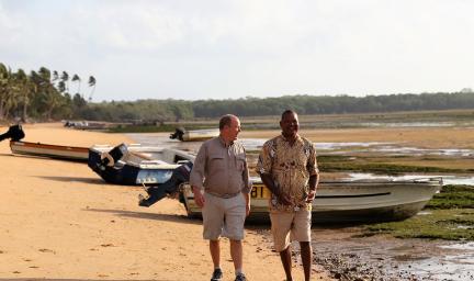 Two men walk along a beach with boats, reef and weed and trees in the background. Both wear shorts and casual shirts.