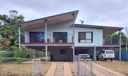 In the foreground are two driveways divided by a wire fence leading to a house divided into two parts and each with two levels. On the right are two vehicles. In the background are trees and a cloudy sky.