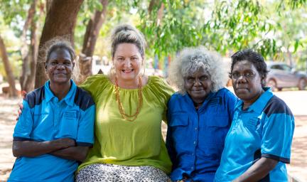 Four women ranging in age from middle-aged to elderly and dressed in different coloured shirts sit in front of trees. In the background are some cars.