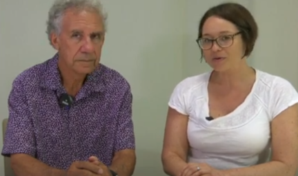 Man and woman set side-by-side at a white desk with a plain grey wall in the background. The man is sat on the left and is wearing a short-sleeve, button-up shirt with a purple pattern. The woman is on the right-hand-side, wearing white t-shirt