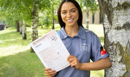 A young woman stands next to a tree holding a certificate in her hands. She is wearing a blue-grey top. In the background are trees and grass and a building.