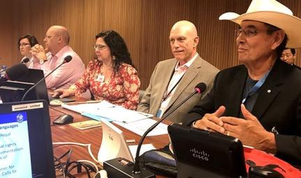 Group of two women and three men sit at a table. Atop the table are microphones and computer terminals. In the background is a timber wall.