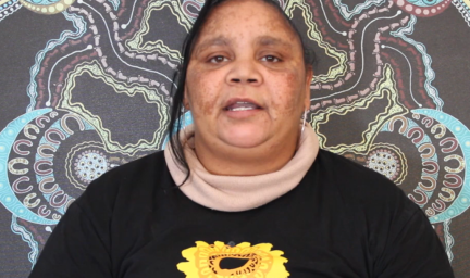 A woman is sitting in front of a blue, black, red and yellow Indigenous artwork. She is wearing a beige turtleneck under a black jumper with a yellow sunflower in the middle.