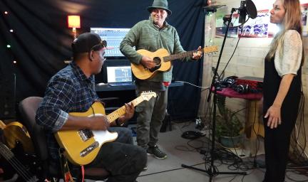 Three people in a small space, wearing casual clothing, are making music. Two men play guitars while a woman stands at a microphone. In the background is recording equipment.