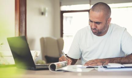 A man in white t-shirt and shaven head sits at a desk writing. In front of him is a laptop, a magazine, papers and booklets. In the background is a wall and windows.