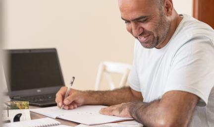 A man smiling while writing in an exercise book seated at a table. Beside him is a laptop and other notebooks.