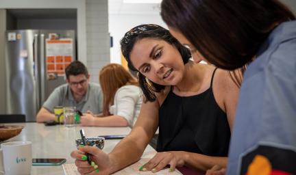 Four young people sitting at a long table look at forms. In the foreground is a young woman in a black top and another in a grey top with the Aboriginal flag on her sleeve. In the background are two other people and a fridge behind them.