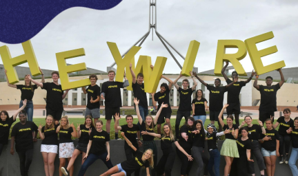 30 young adults are standing in two rows in front of Australian parliament house wearing black t-shirts and smiling. There are 9 young people in the back row holding up the large green letters H E Y W I R E.