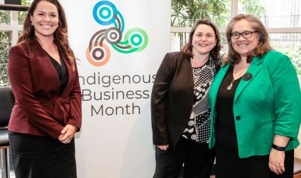 Three women in business wear stand next to a large poster with the words Indigenous Business Month and includes a logo with four circles attached by curved lines. In the background are windows beyond which are trees.